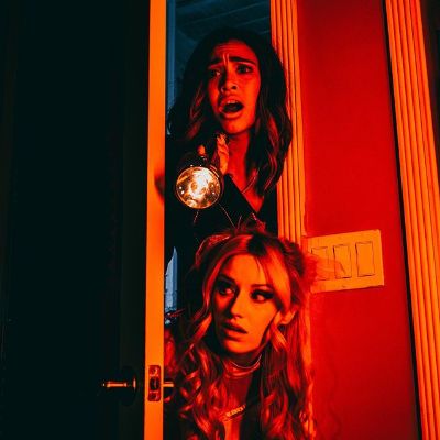 Two scared girls are peeking through the door of a red lit room. 